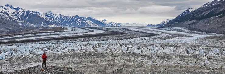 Hiker stands, dwarfed by the glacier, in front of medial moraines snake down to the toe of the south arm of the Lowell Glacier, 8-day packraft expedition down the Jarvis River to the Kaskawulsh River, to the Alsek River, into Lowell Lake, and across to the rock island that splits the Lowell Glacier into two tongues.